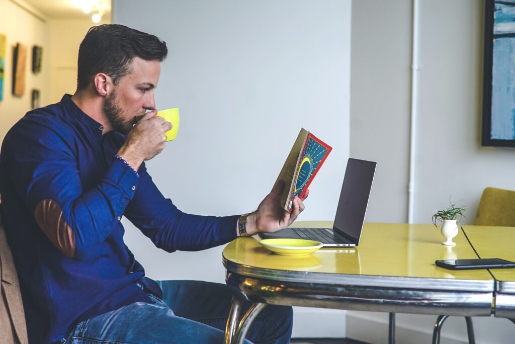 A man is sitting in front of a laptop, reading a book and drinking a hot beverage. Next to it lies his cell phone